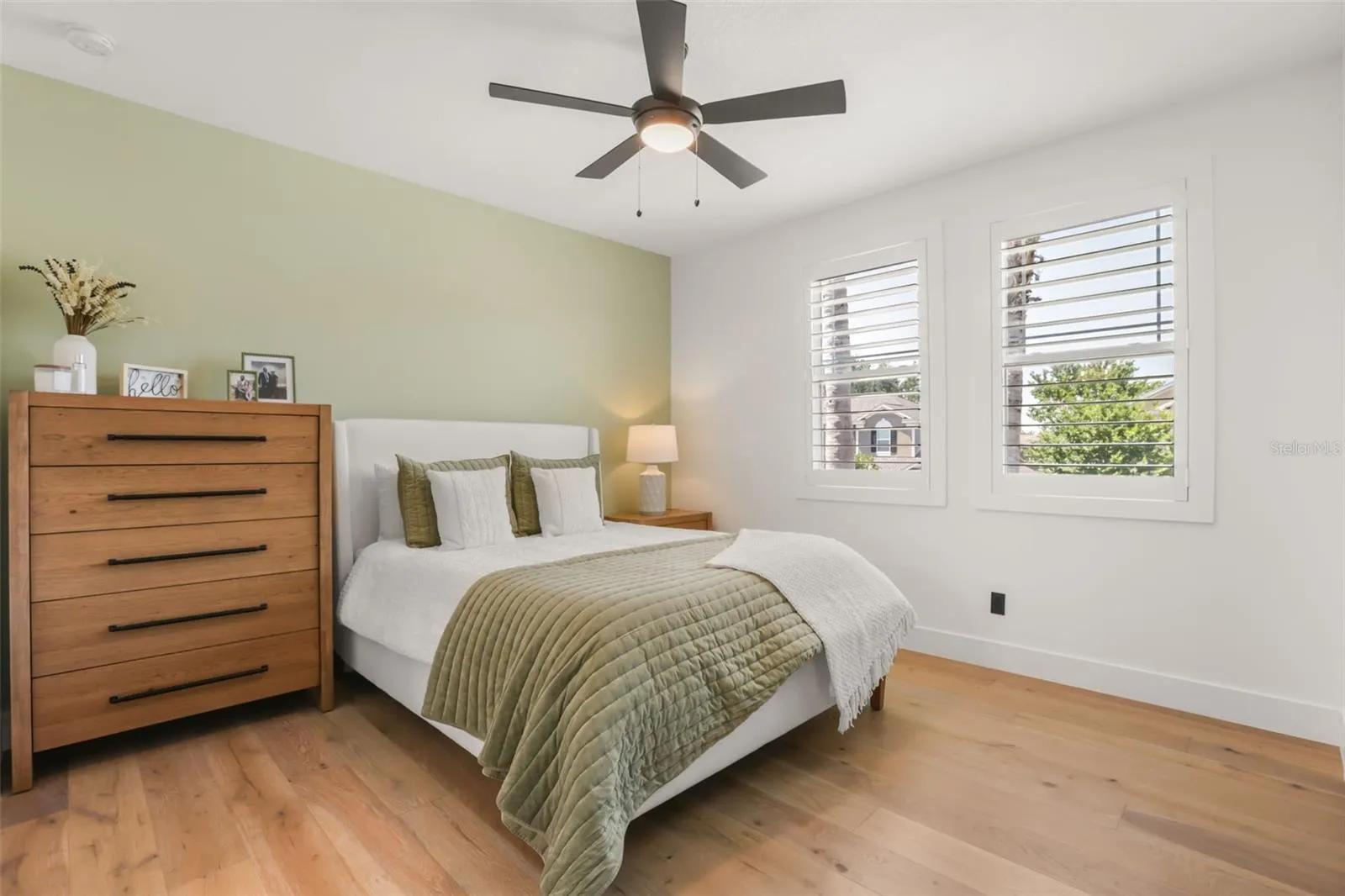 This bedroom features warm wood flooring, plantation shutters, a modern ceiling fan, and a soft green accent wall for a calm, inviting look.