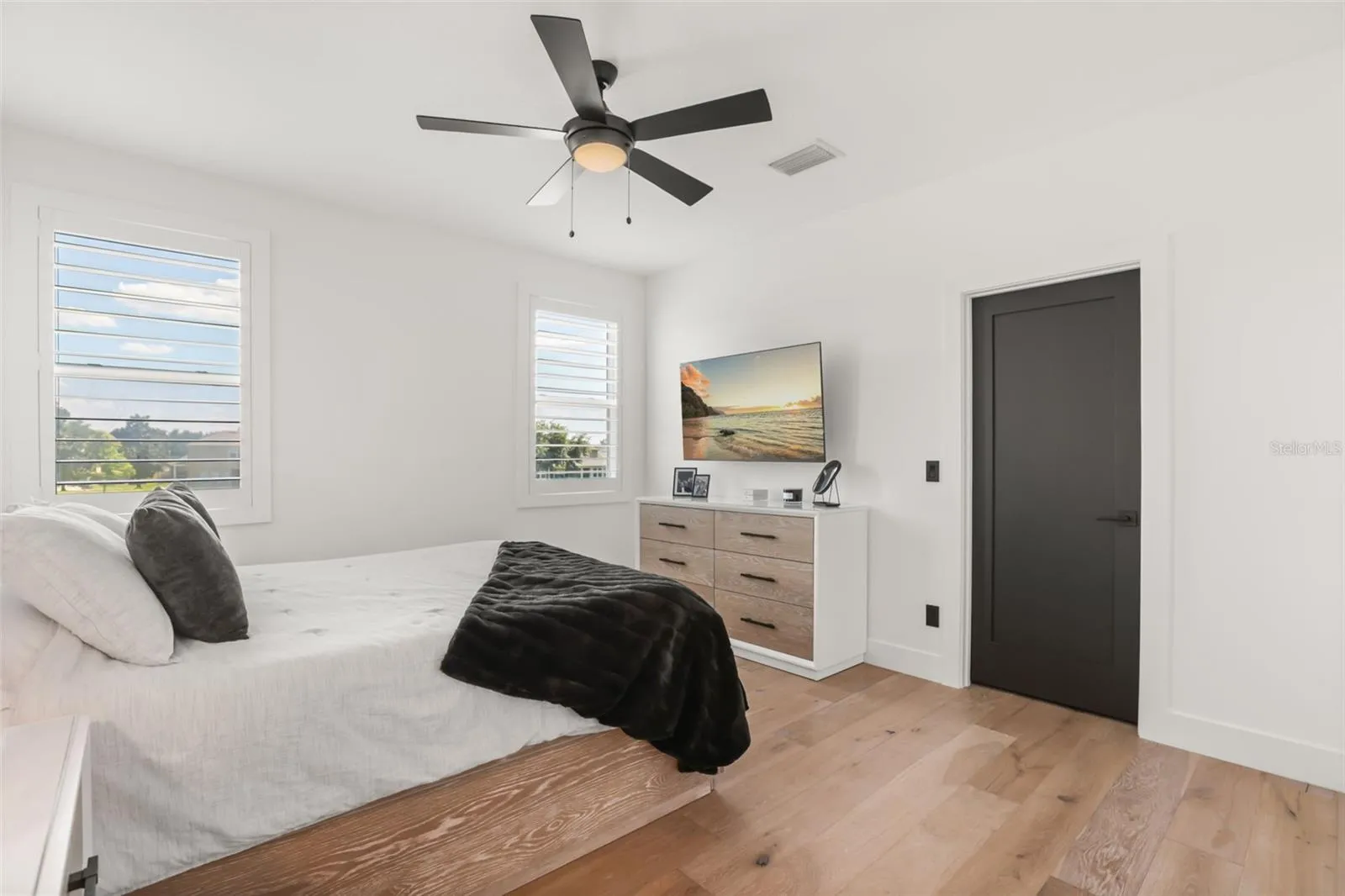 Plantation shutters, wood flooring, and a wall-mounted TV create a comfortable bedroom with a bright, streamlined layout.