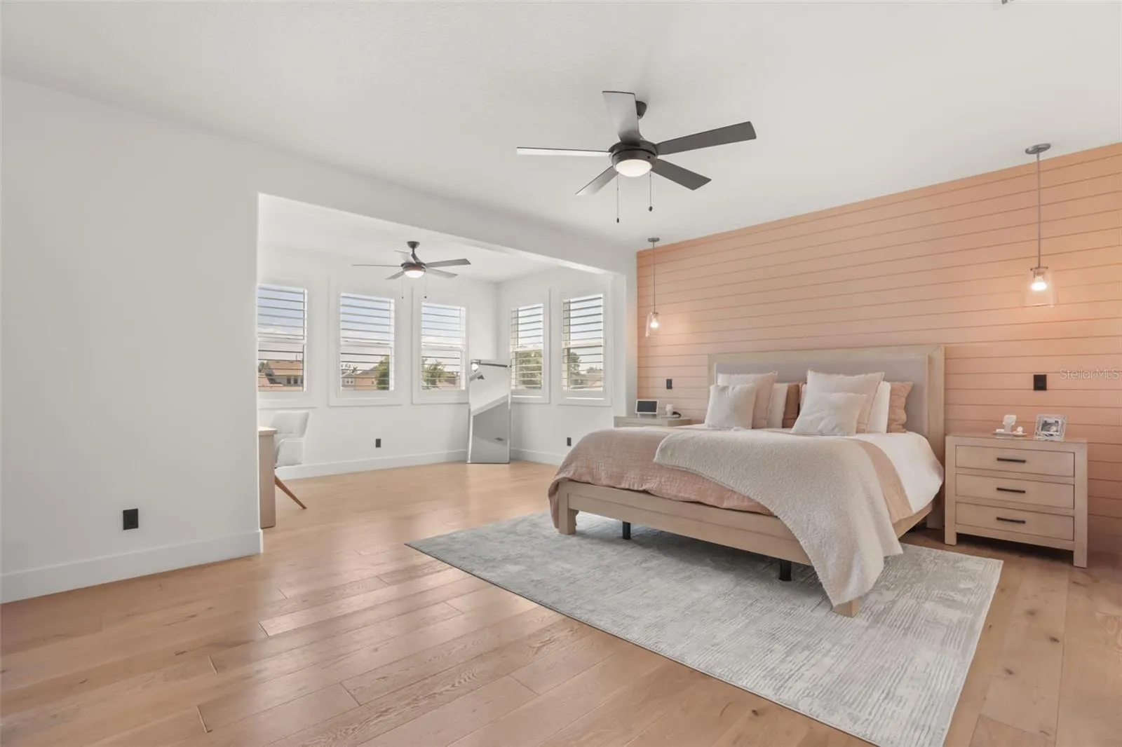 The primary bedroom features warm wood flooring, a soft shiplap accent wall, pendant lighting, and a spacious sitting or vanity area.