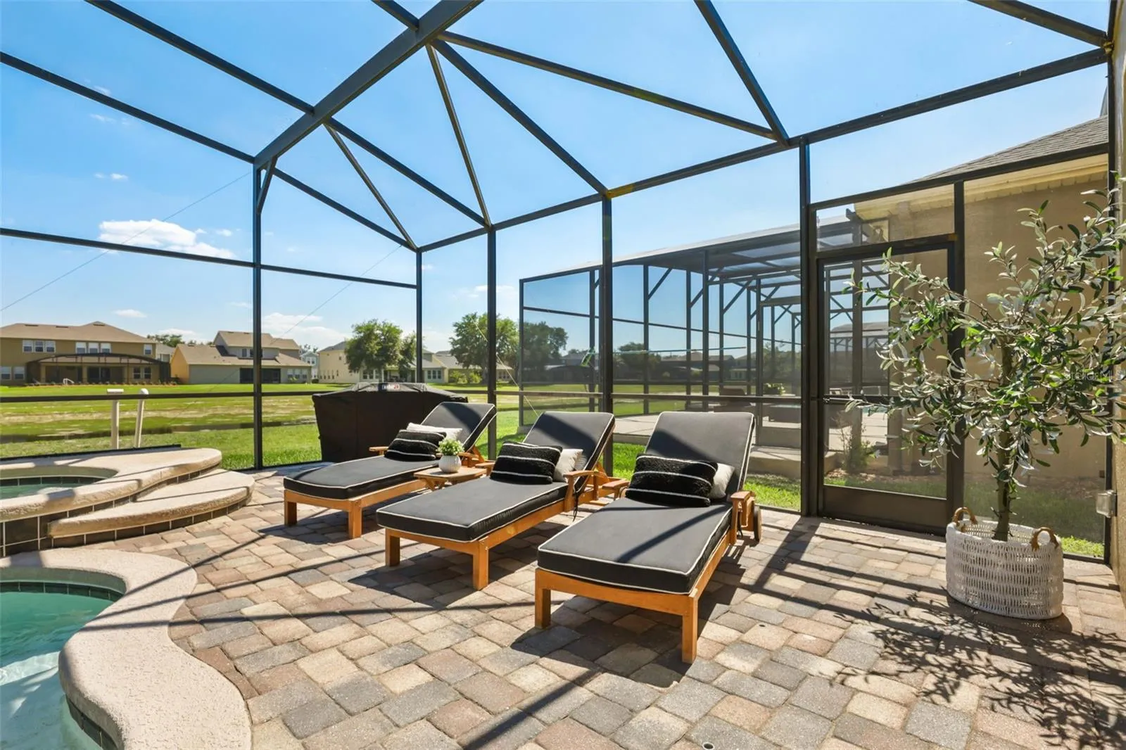 A sunny poolside lounge area features chaise seating, a paver deck, and screened enclosure with open green views beyond.