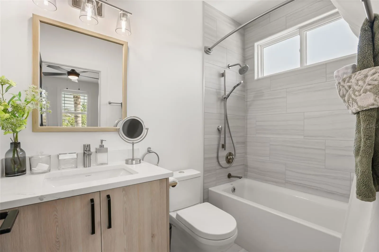 The bathroom includes a tub-shower combination with marble-look tile, a light wood vanity, and a window that brings in natural light.
