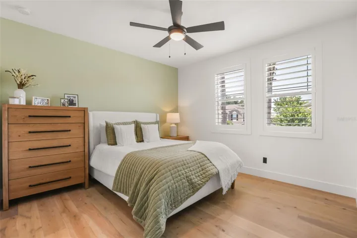 This bedroom features warm wood flooring, plantation shutters, a modern ceiling fan, and a soft green accent wall for a calm, inviting look.