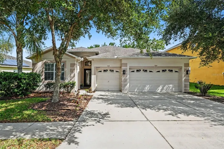 Vew of single-story home with mature tree canopy, lush landscaping, full concrete driveway, and 3-car garage in Live Oak Preserve, Tampa FL 33647