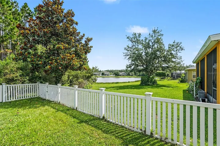 Backyard with white vinyl picket fence, community pond visible beyond fence line, magnolia tree, and screened lanai at right in Tampa FL 33647