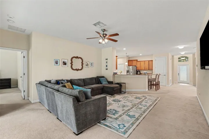 Open-concept family room with ceiling fan, and open sightline to granite kitchen peninsula, bar seating, and foyer in Tampa FL 33647. Primary bedroom suite to left.