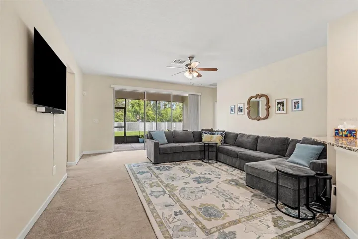 Bright family room with oversized sectional, ceiling fan, and sliding glass door opening directly to screened lanai and backyard in Tampa FL 33647. 2 bedrooms & full bath to the far left, primary suite to the far right