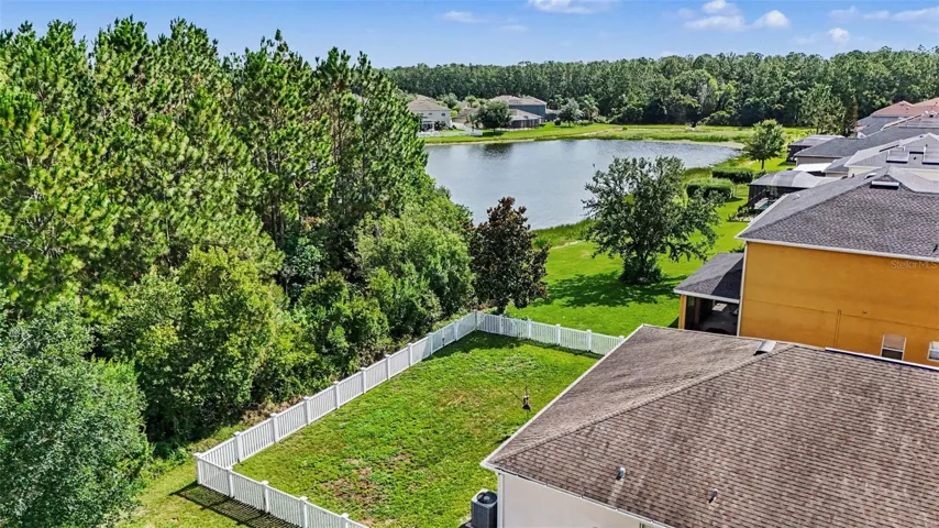 Aerial view of fenced backyard with white vinyl fence, conservation tree buffer, and community pond clearly visible beyond in Tampa FL 33647