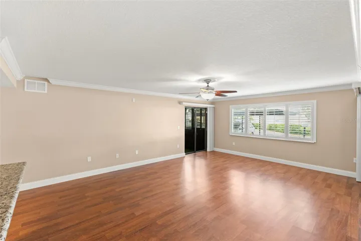 The spacious living room features wood-look flooring, crown molding, plantation shutters, a ceiling fan, and abundant natural light.