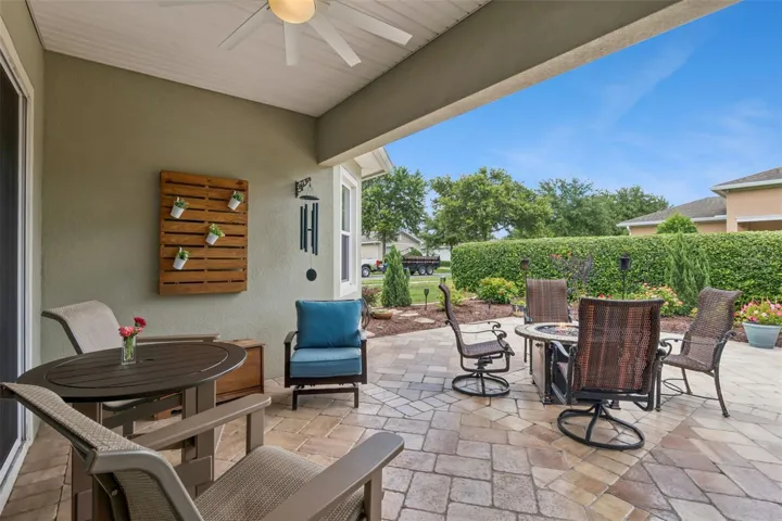 View of the covered lanai w/ ceiling fan, stone paver patio & mature privacy hedge