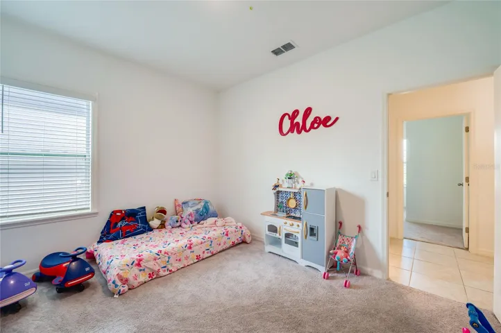 A bright and inviting bedroom layout with light neutral walls, soft carpeting, and an open doorway leading out to a tiled hallway transition.