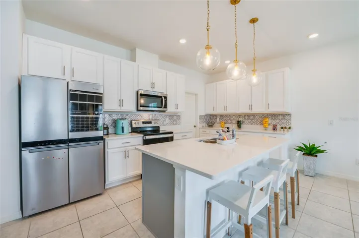 A close-up view of the kitchen showcasing a large island with a double sink, modern glass globe pendant lighting, and ample counter space. The white cabinetry is complemented by sleek stainless steel appliances and a geometric patterned backsplash.