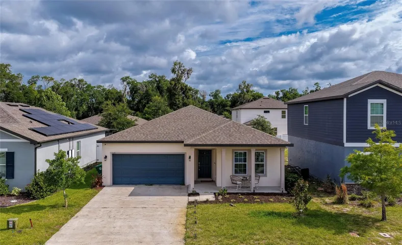 Charming single-story home featuring a two-car garage with a deep blue door, and a welcoming covered front entryway. The property has a concrete driveway and a neatly landscaped front lawn under a bright, cloud-filled sky.