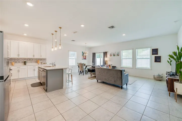 Bright, open-concept main living area showcasing durable tile flooring, recessed ceiling lights, and modern pendant light fixtures. The space seamlessly transitions from a contemporary kitchen with white cabinetry and an island into a spacious living and dining zone lined with large windows.