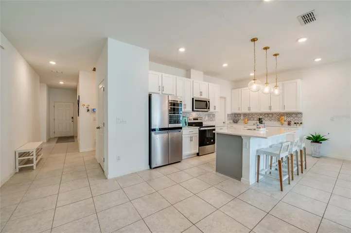 Bright, open-concept kitchen featuring an island with a breakfast bar, white cabinetry, a decorative tile backsplash, and stainless steel appliances. The space flows seamlessly into a tiled hallway leading to the entryway.