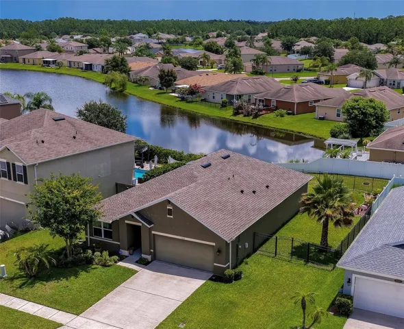 An elevated, angled view of a suburban neighborhood showcasing a single-story home with an attached two-car garage. The property is surrounded by a manicured green lawn and sits near a scenic neighborhood pond.