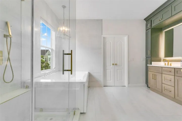 Spa-like bathroom boasts a two-tone vanity with floor-to-ceiling cabinets, a relaxing spa tub, a frameless glass shower, and subtle gray tilework that contrasts beautifully against dark green and light wood cabinetry