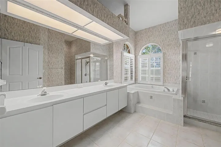 Master bathroom featuring a spacious five-piece layout with an extended dual-sink vanity. The bathroom includes a soaking tub positioned beneath two windows with plantation shutters, providing abundant natural light. A separate walk-in shower and a private toilet area, partially enclosed by a privacy wall, complete this well-designed and functional space.