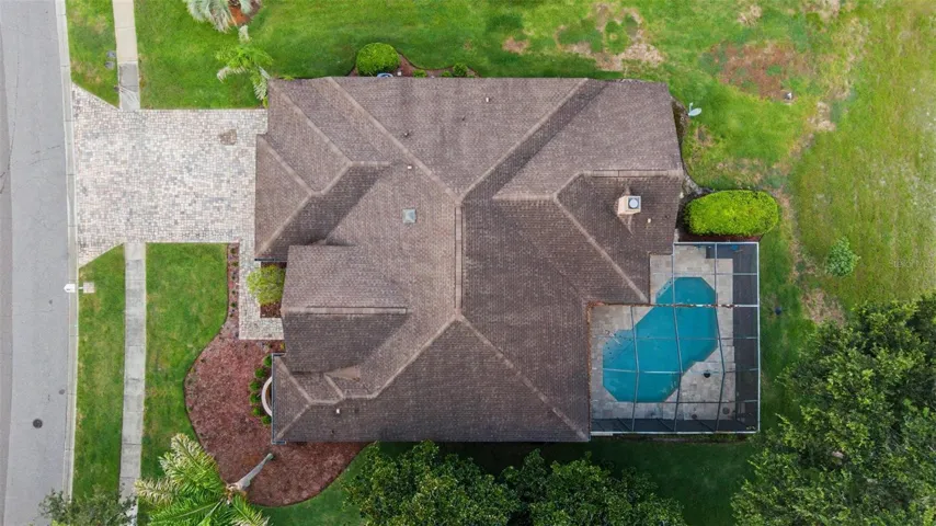 Drone aerial view directly above the home, showcasing the roofline, backyard pool, and surrounding lot. The image highlights the paver driveway, mature landscaping, and overall property layout, offering a clear perspective of the home’s footprint and outdoor living areas.