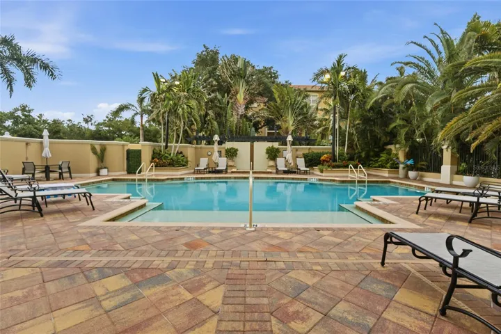 Community pool with sun shelf, spa, and ample chairs on the beautiful paver deck surrounded by lush landscaping