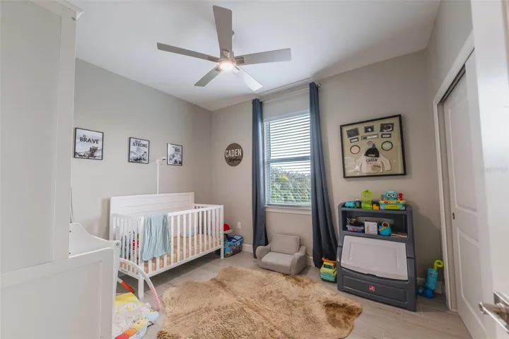 This charming and versatile bedroom is styled as a bright, airy nursery. It features high ceilings, a premium ceiling fan, durable wood-look flooring, and a large window providing beautiful backyard views, illustrating the highly flexible layout of this custom home.