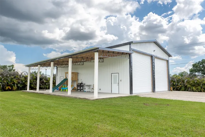 The ultimate hobbyist setup! This massive detached steel workshop/garage features dual high-clearance roll-up doors and a huge covered lean-to/patio area—perfect for an outdoor kitchen, boat storage, or a massive covered play space.
