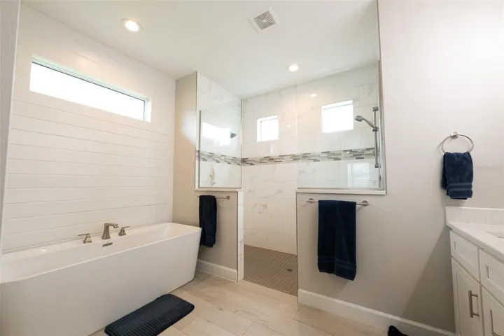 Escape to your private spa! The luxurious primary bathroom suite features a freestanding soaking tub set against a modern white shiplap accent wall, high-end wood-look porcelain tile flooring, and a massive, custom walk-through double shower lined with marble-style tile.