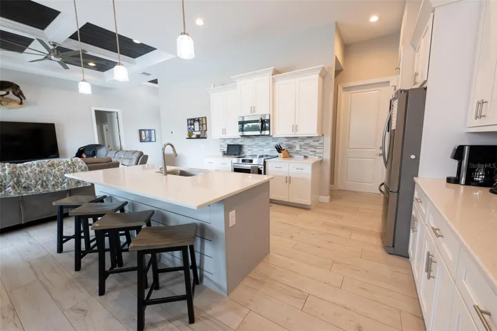 This wide-angle view from the kitchen showcases the highly desirable open-concept floor plan. Chat with guests at the island bar while overlooking the living room and taking in the architectural beauty of the custom ceilings and uniform wood-look tile floors.
