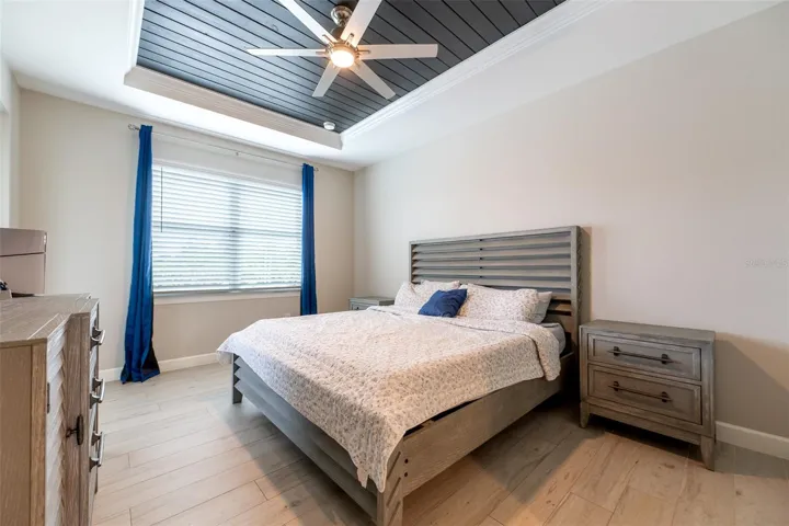 The expansive primary bedroom serves as a true sanctuary, highlighting a dramatic recessed tray ceiling inlaid with dark wood paneling and a modern white ceiling fan. Large windows frame beautiful outdoor views and flood the room with natural light.