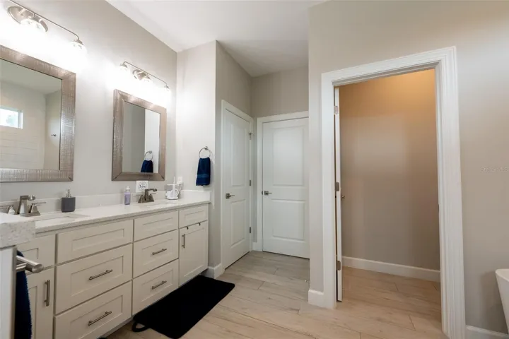 Reverse view of the primary vanity showing the seamless flow of the master suite. High ceilings, uniform wood-look tile flooring, and clean neutral tones lead directly to a private water closet and the expansive walk-in master wardrobe.