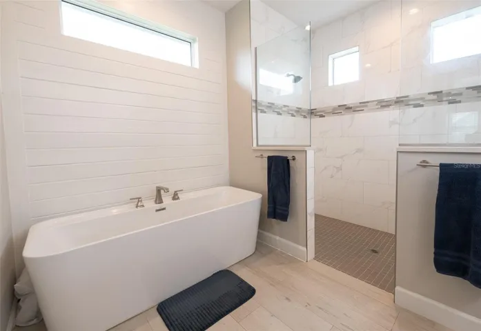 A closer look at the stunning freestanding tub and walk-through shower layout. The massive open-concept shower boasts custom marble-patterned porcelain wall tile, a linear mosaic accent borders, dual entryways, and premium fixtures.