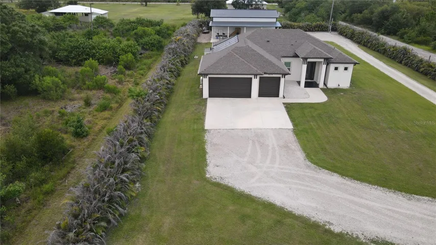 High-altitude drone overview illustrating the true scale and impressive privacy of this custom compound. Lined completely by dense, mature tropical privacy hedges, the estate boasts a grand gravel driveway turnaround, expansive cleared green spaces, and a perfect configuration between the primary residence and the massive detached outbuilding workshop.