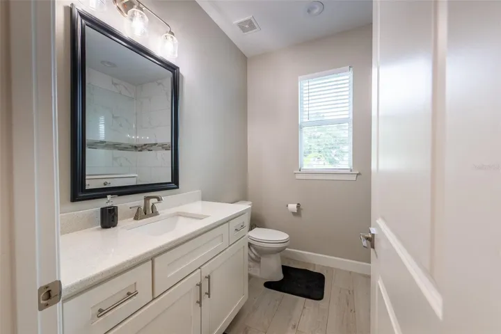 This pristine guest bathroom is loaded with premium finishes, boasting an oversized single vanity with custom white shaker cabinetry, elegant brushed nickel hardware, a sparkling quartz countertop, uniform tile flooring, and a bright window.
