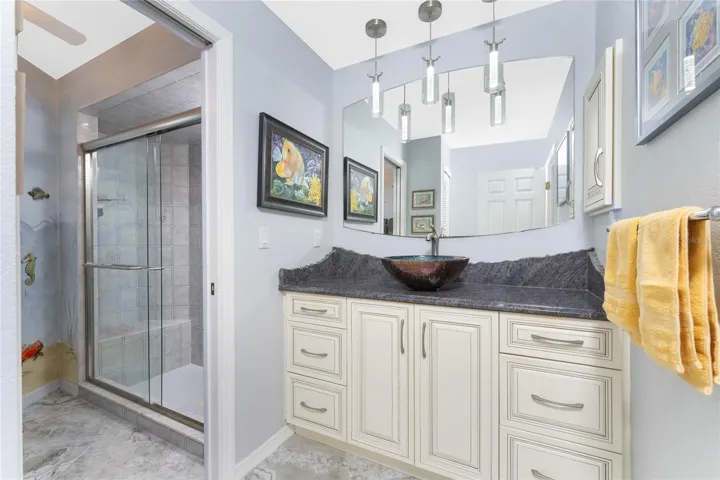 This bathroom showcases a stunning vessel sink atop elegant gray granite countertops, solid wood soft close white cabinetry, modern pendant lighting, and a walk-in shower."