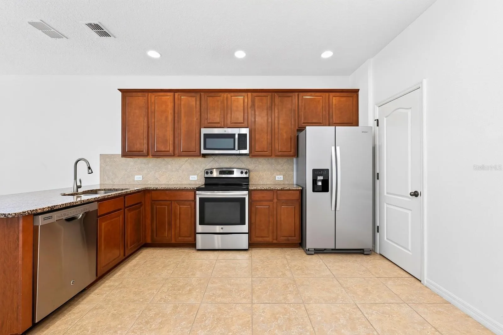 The kitchen is designed to be both functional and inviting, featuring rich cabinetry, granite countertops, a tiled backsplash, STAINLESS STEEL APPLIANCES, pantry storage, and a breakfast bar that overlooks the living and dining areas.