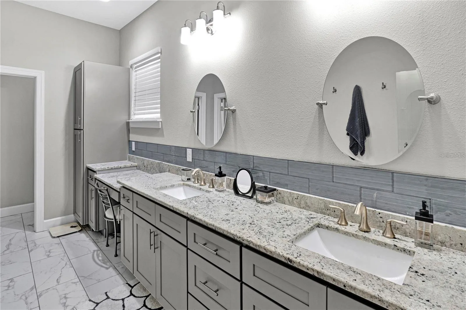 The luxurious primary bathroom features an extended double vanity with granite tops, a dedicated makeup vanity station, contemporary gray cabinetry, and a spacious walk-in shower