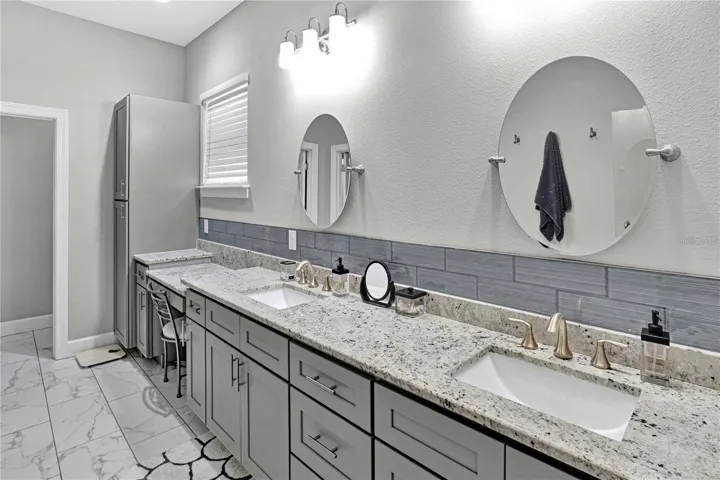 The luxurious primary bathroom features an extended double vanity with granite tops, a dedicated makeup vanity station, contemporary gray cabinetry, and a spacious walk-in shower