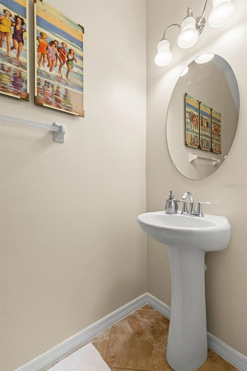 The first-floor powder room features a pedestal sink, oval mirror, down light fixture and neutral tile flooring.