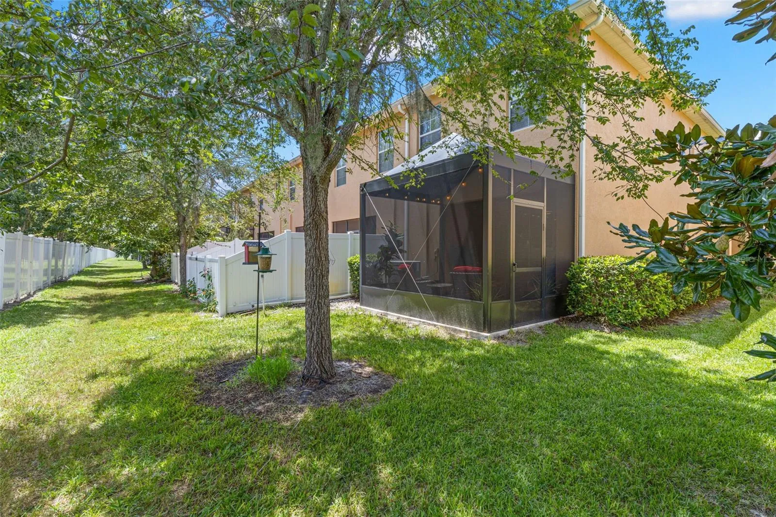From the rear, the 2026 screened lanai sits beneath a canopy of mature trees, with a grassy common area adding space and greenery beyond.