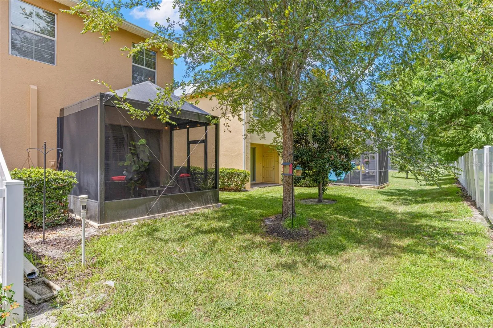 The rear common area is lined with mature trees and a vinyl fence, creating a green, shaded backdrop behind the screened lanai.