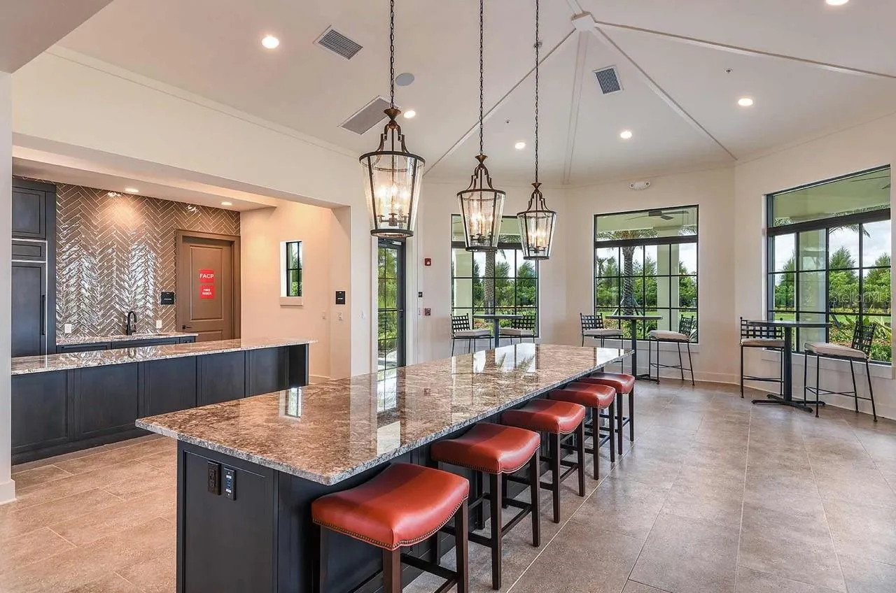 Vaulted, beamed ceilings and a wall of windows fill the clubhouse's social kitchen and gathering room with natural light, anchored by a long stone-topped island with bar seating. Bistro tables near the windows offer additional spots to linger over coffee or catch up with neighbors.