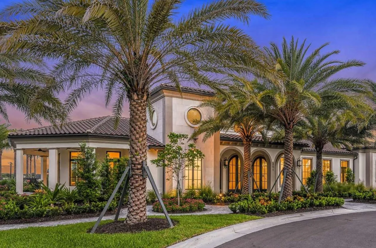 Warm interior lighting glows through arched, multi-pane windows as dusk settles over the clubhouse facade, framed by mature palms and a paver motor court. Stone accents, a tile roofline, and covered walkways give the building a welcoming, gathering-place feel from the moment residents arrive.