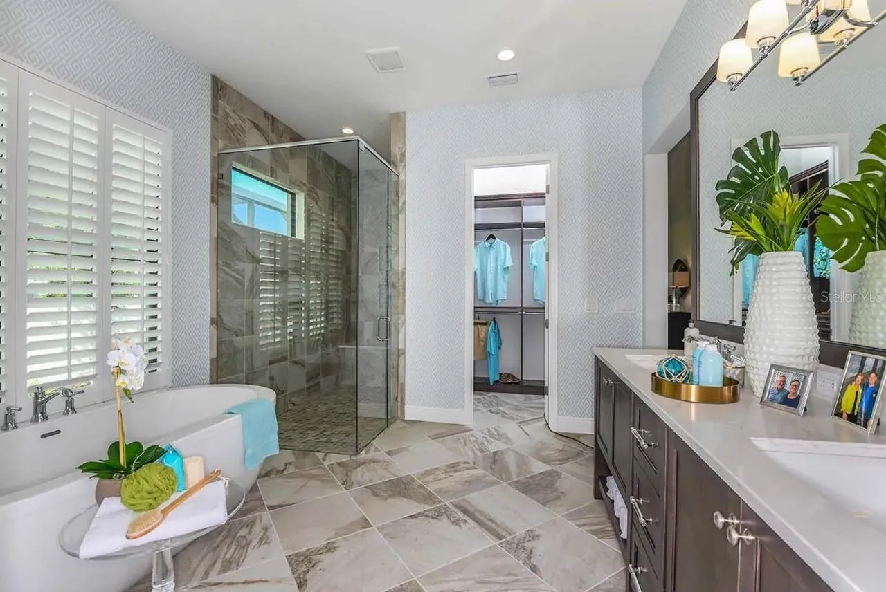 A freestanding soaking tub sits beneath a shuttered window, paired with a glass-enclosed shower, marble-look tile flooring, and a furniture-style vanity with dual sinks. A walk-in closet is visible just beyond, keeping the suite's layout efficient.DISCLAIMER: Images shown are of a furnished model and are for illustration purposes only. Images are not an exact representation of this feature home and this home will not be sold furnished