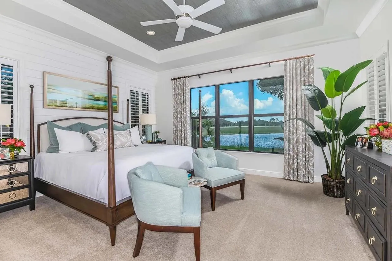 A four-poster bed anchors this bedroom, finished with a tray ceiling, patterned drapery, and a window seating area that takes in the water view outside. Soft teal accents throughout add a calming, coordinated palette.DISCLAIMER: Images shown are of a furnished model and are for illustration purposes only. Images are not an exact representation of this feature home and this home will not be sold furnished