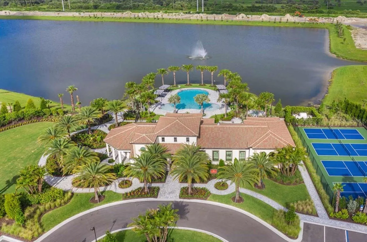An aerial view frames Aria's clubhouse alongside a curving lake and its working fountain, with lighted tennis and pickleball courts, a resort-style pool, and palm-lined walkways below. The scale of the grounds offers a first look at the everyday resort feel awaiting residents just beyond their own front door.