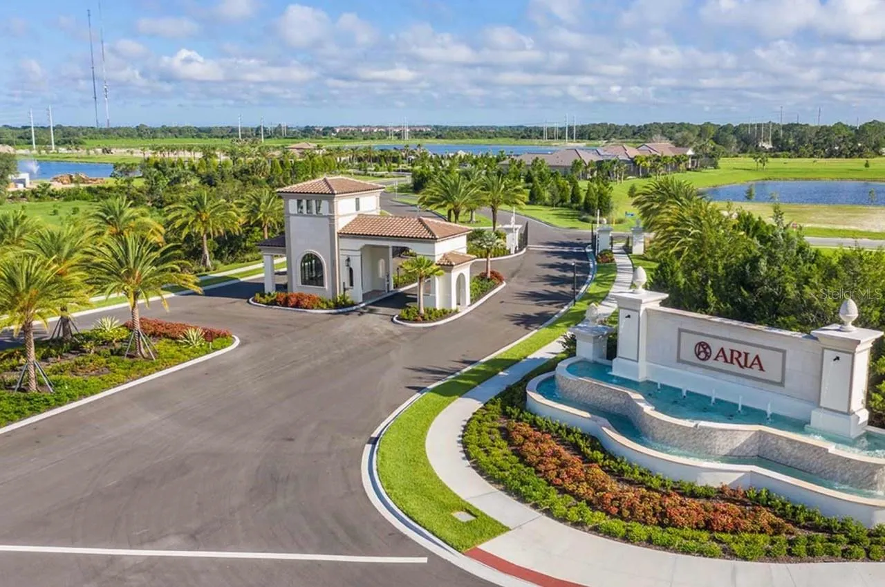 An aerial perspective takes in Aria's full entry sequence, from the fountain-flanked monument sign to a covered gatehouse-style structure and winding entry drive, framed by lakes and lush landscaping. The layout offers a clear sense of the community's scale and setting.