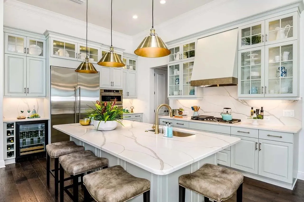 A marble-look waterfall island anchors this kitchen, paired with glass-front cabinetry, a full-height tile backsplash, and a row of brass pendant lighting overhead. A built-in wine fridge and stainless appliances round out the space.DISCLAIMER: Images shown are of a furnished model and are for illustration purposes only. Images are not an exact representation of this feature home and this home will not be sold furnished.