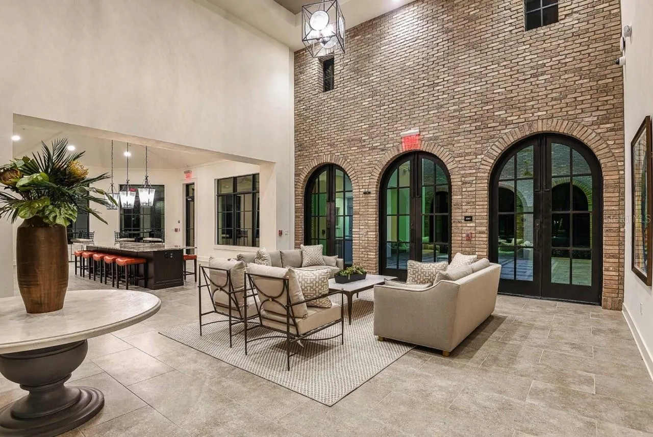 An arched, brick-columned loggia opens onto a shaded outdoor seating area, furnished for residents to relax or socialize before or after using the clubhouse. Wood-beamed ceilings and pendant lighting carry the same finishes found throughout the clubhouse into this open-air gathering spot.