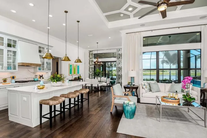 An open kitchen and living space is finished with a wood-look tile floor, bronze pendant lighting, and a wall of glass sliders that frame a water and palm view beyond. A patterned area rug and soft seating create a defined gathering spot just steps from the island.DISCLAIMER: Images shown are of a furnished model and are for illustration purposes only. Images are not an exact representation of this feature home and this home will not be sold furnished