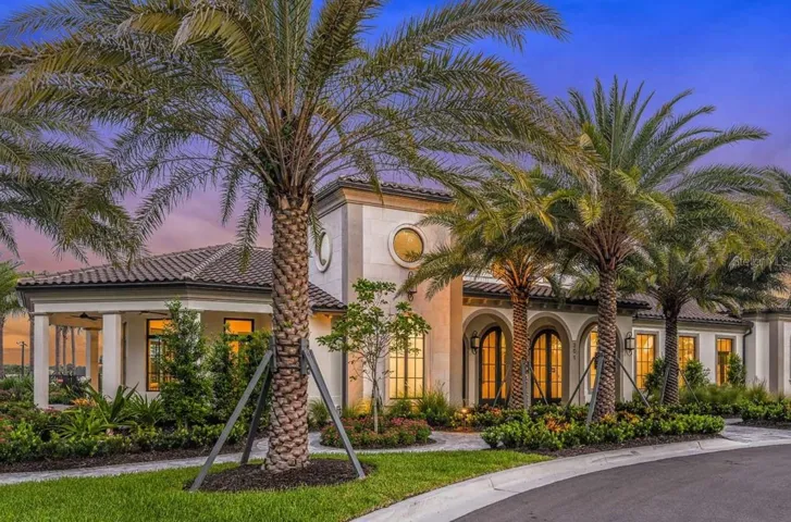 Warm interior lighting glows through arched, multi-pane windows as dusk settles over the clubhouse facade, framed by mature palms and a paver motor court. Stone accents, a tile roofline, and covered walkways give the building a welcoming, gathering-place feel from the moment residents arrive.