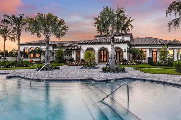 As the sky turns pink and gold, the clubhouse pool and sundeck glow beneath market lighting, with the building's arched entry and tile roof visible just beyond the water's edge. Wide paver decking and in-pool handrails complete a resort-caliber setting for evening swims.